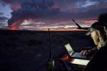 Two caucasian men's faces illuminated by the glow of a laptop screen with a glowing pink sunset and fire on the horizon