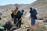 Three people stand at bottom of hill facing away from camera. Person in middle holds a hose spraying water. 