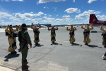 A group of people standing on airplane tarmac wearing tan jumpsuits and black gear strapped to their front look up with their hands raised.