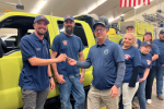 Caucasian men and women pose in front of a yellow truck, two people exchanging a set of keys