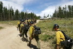 Caucasian men in yellow shirts, black packs, and black hardhats walk in single file on a dirt road.