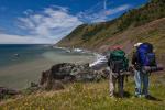 Two people wearing hiking backpacks look out at the beach below a hill.