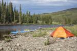 A peaceful, dark river snakes through the middle of the photo with a rocky bank in the foreground and thick forest, hills, and cloudy sky in the background. There’s an orange tent on the rocks in the lower right of the image and two blue kayaks perched on the shoreline a few feet away. A man squats in the middle of the image with his back to the river near the kayaks. 