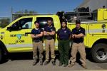 A crew of four firefighters stand in front of their fire engine.