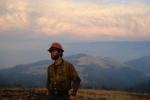 a firefighter wearing a hard hat and Nomex poses in front of a smokey landscape of hills