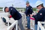 Three workers wearing hard hats and navy shirts conduct an inspection on industrial equipment outdoors with green fields in the background.