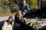 Three people are crouched by a pickup truck, displaying a row of captured iguanas on the bed, surrounded by greenery.