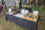 Outdoor display table with antique diving helmet, boots, weights, nautical tools, maps, and informational signs at a historical event.