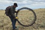 A man has a large old barbed wire fence in his hand in a large circle with clouds in the sky and brown grass. 