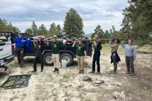 Seven people in a row holding a long pole on their shoulders. Trailer full of trash behind them. 