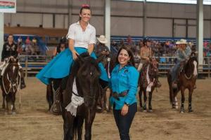 A person in a blue shirt and blue skirt sits on top of a dark brown horse and a person in a blue shirt and black pants stands next to them.  